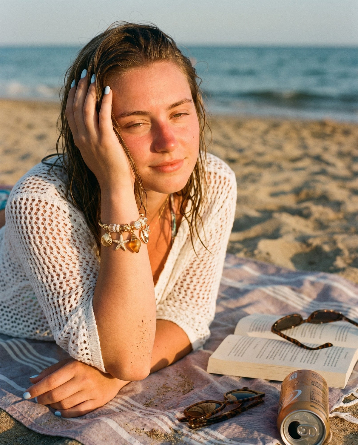 Young woman on a beach towel at golden hour wearing a gold seashell charm bangle