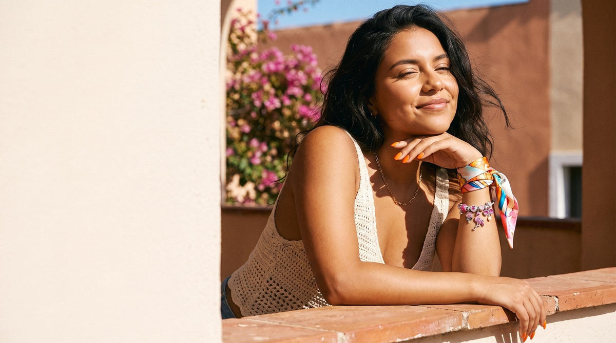Young woman leaning on a sunlit balcony rail wearing a gold charm bangle