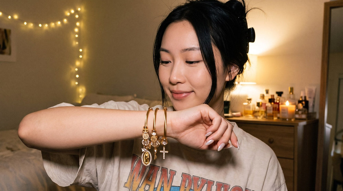 Young woman checking three stacked gold charm bangles on her wrist in a warm-lit bedroom